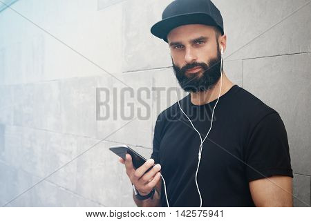 Bearded Muscular Man Wearing Black Tshirt Blank Snapback Cap Summer Time.Young Men Smiling Opposite Empty Gray Concrete Wall Background Using Smartphone Headphones.Horizontal Mockup.Side View