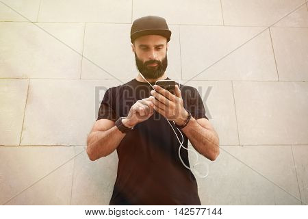 Bearded Muscular Man Wearing Black Tshirt Blank Snapback Cap Summer Time.Young Men Standing Opposite Empty Gray Concrete Wall Background Using Smartphone Headphones.Horizontal Mockup.Color Filter