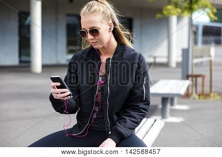 Beautiful woman with sunglasses sits on a bench in the city wearing a jacket and sportswear. Listens to music and using smart phone.