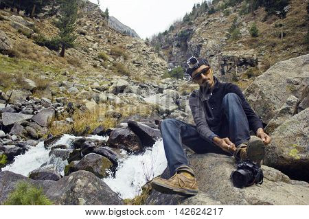Young photographer Aleem Zahid Khan hiking Sitting on rock and tying his shoes in the mountains near waterfall