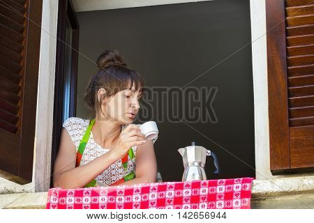 Portrait of concentrated woman is looking at hot coffee cup in her hand sitting near open window with traditional European wood brown shutters