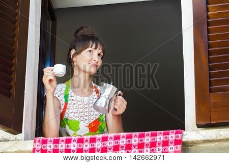 Portrait of happy woman is enjoying hot coffee drink freshly brewed in Italian moka pot sitting near open window with traditional European wood brown shutters