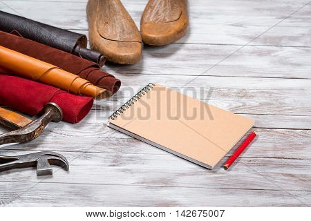 Brightly colored leather in rolls working tools shoe lasts notebook with pencil on white wooden background. Leather craft.