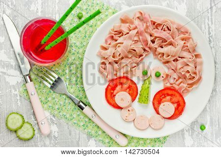 Breakfast for child. Pink noodles with sausage tomato cucumber and green peas in the form of scary man face. Concept healthy and creative baby food top view