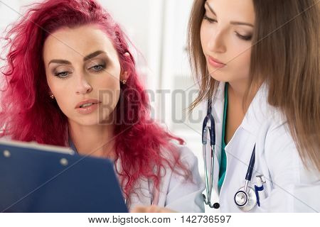 Two female doctors looking at clipboard with patient information form discussing his diagnosis and treatment. Health care medical education and service concept.