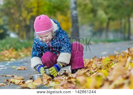 Two years old girl is holding thermos cup hunkering down in autumn foliage covering backdrop