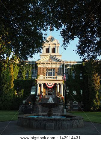 The old Crook County Courthouse in Prineville in Central Oregon is beautifully lit as the sun goes down. The railings on the steps have flower baskets the American flag flies at half-mast and the fountain flows.