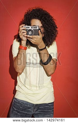 Closeup of one handsome passionate expressive cool young brunette photographer men with long curly hair holding a vintage SLR camera standing against red background