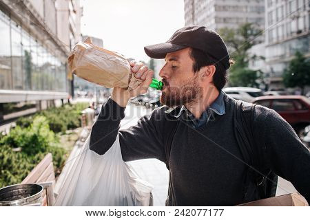 A Picture Of Man Standing On The Street And Drinking From Bottle. He Is Homeless. Guy Has A Bag Hang