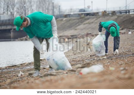 Young men protecting environment by picking up litter from coast by riverside