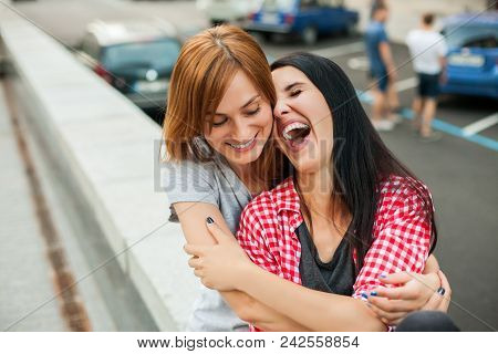 Two Young Teenagers Hugging, Laughting And Sitting Near Road In The Street. One Brunette Girl In Red
