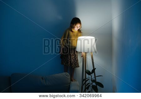 Little Girl In Sweater Standing On A Couch Looking At Nigth Light At Home.