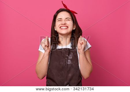 Charming Brunette Female Dresses Brown Apron Dirty With Flour, Expresses Happyness, Clenching Her Fi