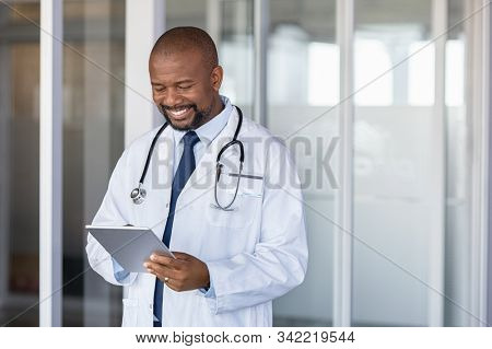 Mature african doctor using digital tablet in corridor . Portrait of confident male doctor using tablet computer in clinic with copy space. Successful smiling doctor in labcoat wearing stethoscope.