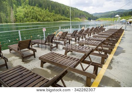 Row Of Empty Wooden Beach Chairs Near Water.