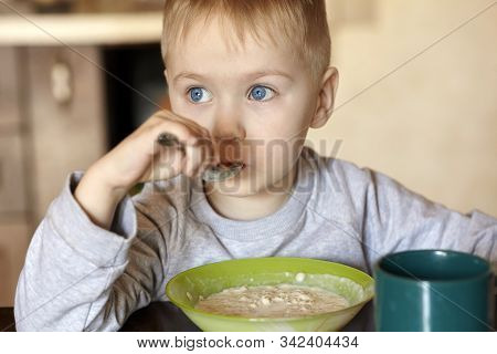 Cute Caucasian Baby Boy With Blue Eyes And Blond Hairs Eating Porridge. Serious Thoughtful Look, Chi