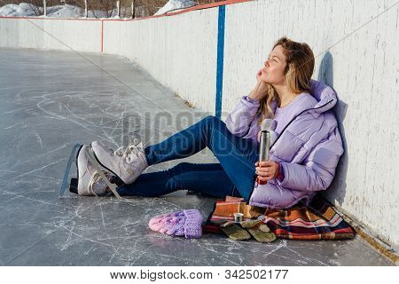Lovely Young Woman Relaxing After Riding Ice Skates And Drinking Hot Drink From Termo Pot On The Ice