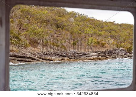 Rocky Coastal Shoreline With Brown Layered Volcanic Shale And Spindly Trees And Sparse Vegetation, A