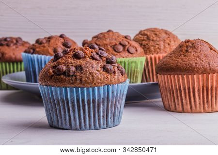 A Few Chocolate-sprinkled Muffins In Colored Cups On A White Wooden Surface. In The Background, Muff