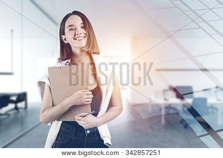 Portrait Of Smiling Young Woman With Fair Hair In Casual Clothes Holding Clipboard Standing In Blurr