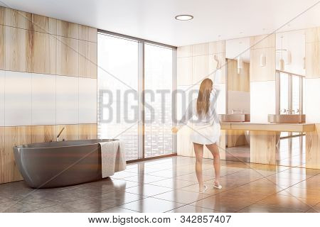 Rear View Of Young Woman Standing In Stylish Sunlit Bathroom With White And Wooden Walls, Tiled Floo