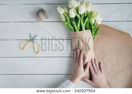 Woman Florist Wrapping Beautiful Bouquet Of White Tulips In Pack Craft Paper On The Wooden Table. Fl