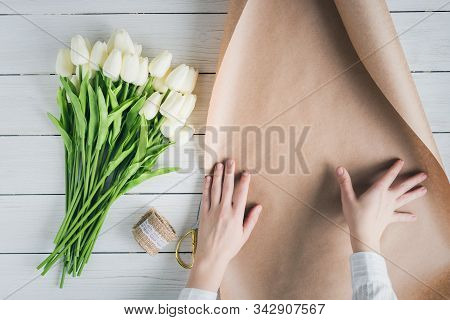 Woman Florist Wrapping Beautiful Bouquet Of White Tulips In Pack Craft Paper On The Wooden Table. Fl