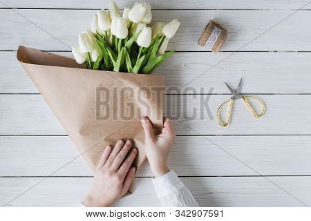 Woman Florist Wrapping Beautiful Bouquet Of White Tulips In Pack Craft Paper On The Wooden Table. Fl