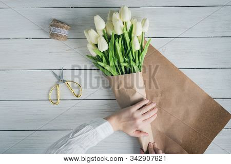 Woman Florist Wrapping Beautiful Bouquet Of White Tulips In Pack Craft Paper On The Wooden Table. Fl