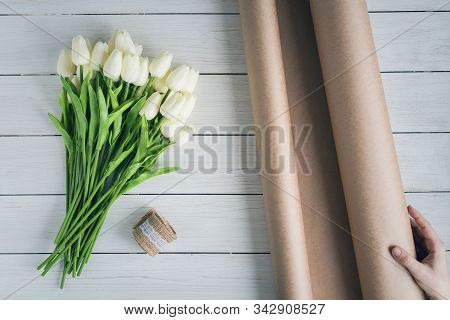 Woman Florist Wrapping Beautiful Bouquet Of White Tulips In Pack Craft Paper On The Wooden Table. Fl