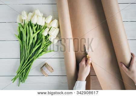 Woman Florist Wrapping Beautiful Bouquet Of White Tulips In Pack Craft Paper On The Wooden Table. Fl