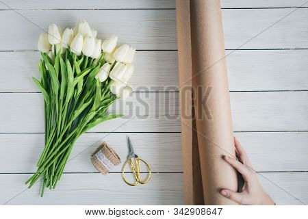 Woman Florist Wrapping Beautiful Bouquet Of White Tulips In Pack Craft Paper On The Wooden Table. Fl