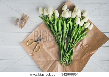 Woman Florist Wrapping Beautiful Bouquet Of White Tulips In Pack Craft Paper On The Wooden Table. Fl
