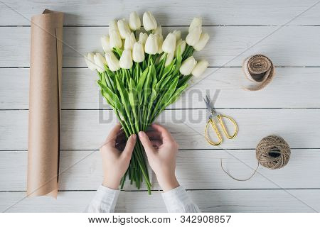 Woman Florist Wrapping Beautiful Bouquet Of White Tulips In Pack Craft Paper On The Wooden Table. Fl