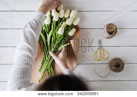 Woman Florist Wrapping Beautiful Bouquet Of White Tulips In Pack Craft Paper On The Wooden Table. Fl