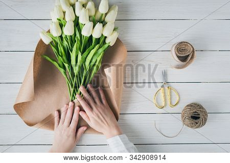 Woman Florist Wrapping Beautiful Bouquet Of White Tulips In Pack Craft Paper On The Wooden Table. Fl
