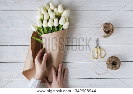 Woman Florist Wrapping Beautiful Bouquet Of White Tulips In Pack Craft Paper On The Wooden Table. Fl