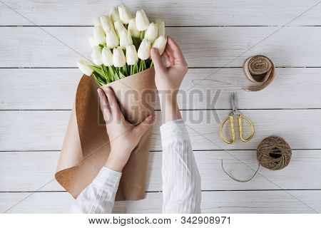 Woman Florist Wrapping Beautiful Bouquet Of White Tulips In Pack Craft Paper On The Wooden Table. Fl