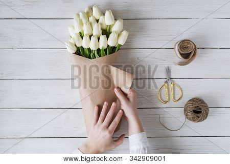 Woman Florist Wrapping Beautiful Bouquet Of White Tulips In Pack Craft Paper On The Wooden Table. Fl