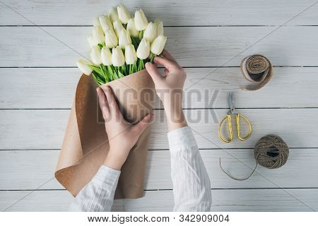 Woman Florist Wrapping Beautiful Bouquet Of White Tulips In Pack Craft Paper On The Wooden Table. Fl