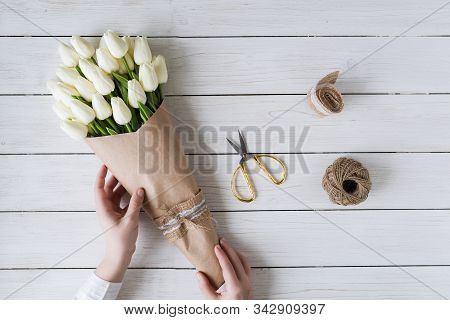 Woman Florist Wrapping Beautiful Bouquet Of White Tulips In Pack Craft Paper On The Wooden Table. Fl