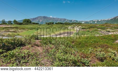 Green Grass And Shrubs Grow In The Crater Of An Extinct Volcano. A Picturesque Mountain Range Agains