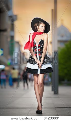 Fashionable lady wearing elegant dress, black hat and red scarf posing outdoor in urban scenery