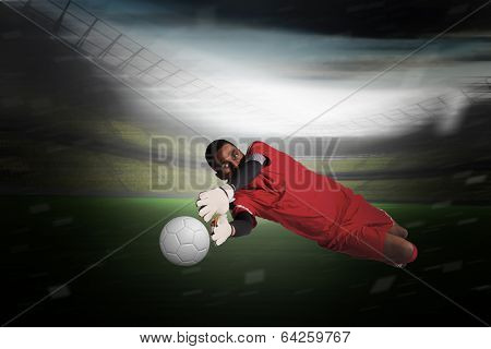 Goalkeeper in red making a save in a large football stadium with lights