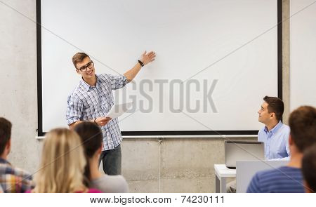 education, high school, technology and people concept - student standing with remote control, laptop computer in front of teacher and classmates in classroom