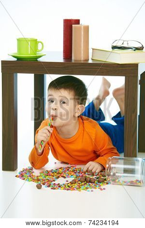 Happy kid eating lollipop under table sweets spilt. Home indoor, isolated on white.