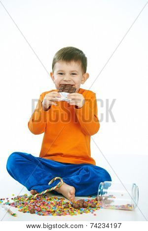 Joyous little kid eating chocolate sitting cross-legged on floor, sweets spilt. Isolated on white.