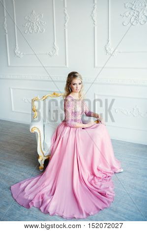 godlike blonde girl in pink wedding dress sitting in the chair in classic white interior.