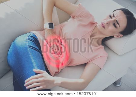 Painful rest. Stressed pretty woman suffering from stomachache and putting arm on her belly while lying on the sofa.