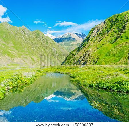 The green slopes of Sno gorge and snowy mountains on the distance reflect in waters of the tiny pond Kazbegi Georgia.